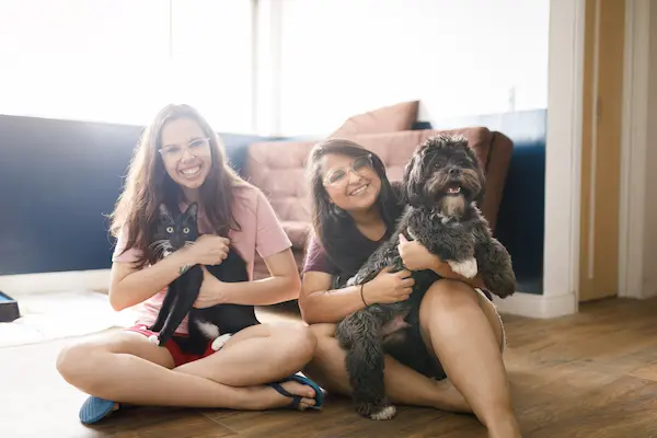 Two women smiling and sitting on hardwood floor with cat and dog.