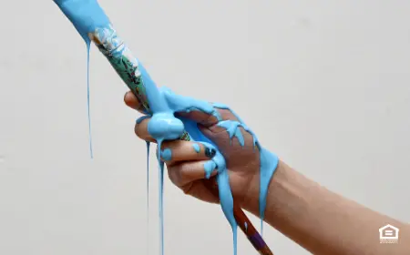 Hand holding paintbrush with blue paint dripping all over.