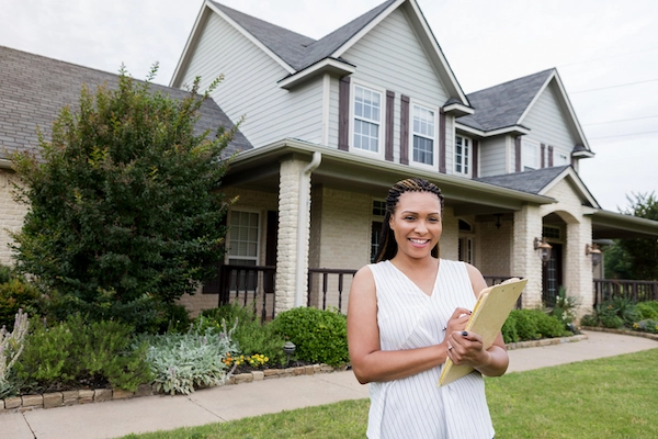 Smiling African American woman holding clipboard standing outside modern two story house.