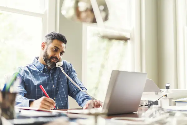 Man sitting in front of laptop holding red pen and corded phone.