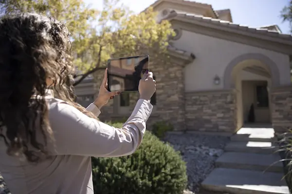Woman holding up tablet device to photograph sunny exterior of residential home.